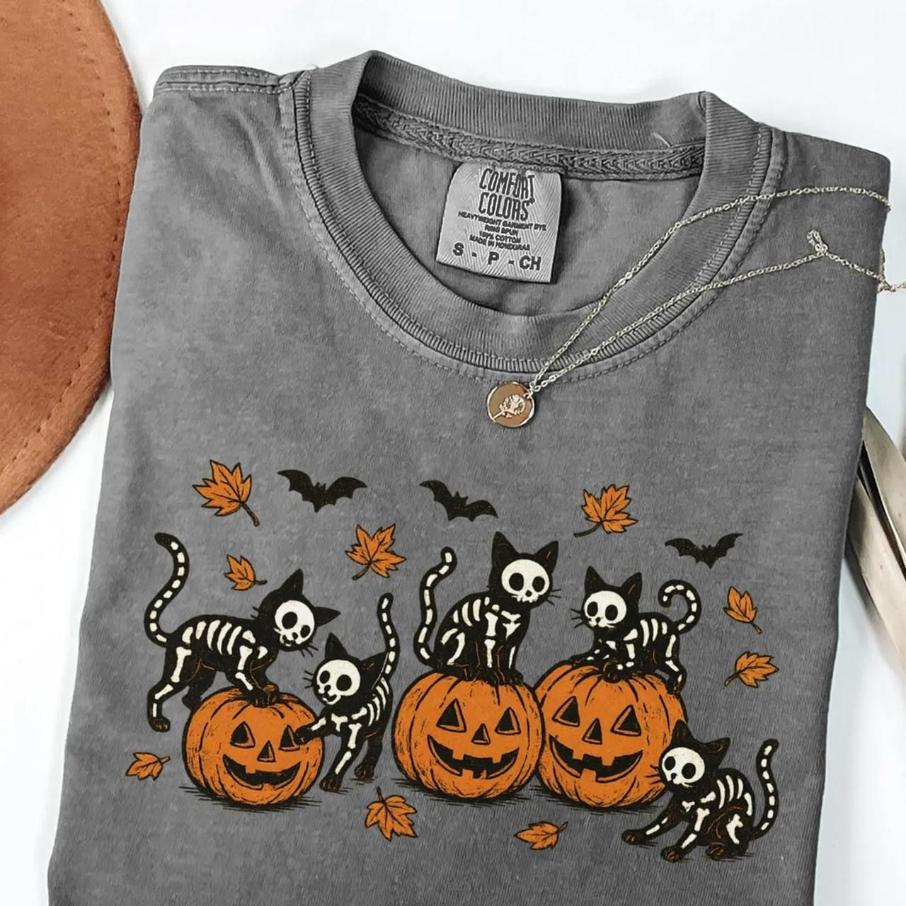 A gray t-shirt with a Halloween design featuring skeletal cats on carved pumpkins, surrounded by flying bats and falling autumn leaves.